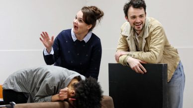 Three people interact indoors; one person leans forward, head down on a sofa, hiding their face, while another stands behind, smiling and waving, and a third leans on a prop, also smiling.