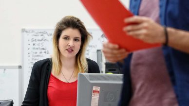A woman sits at a desk with a computer, looking at a man in the foreground who is holding a red folder and standing. A whiteboard with writing is in the background.