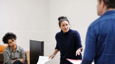 Three people in a rehearsal room: a woman in a dark jumper gestures passionately whilst holding a script, a seated woman watches attentively, and a man in a blue shirt stands facing them, partially out of view.