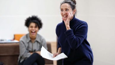 A woman in a dark jumper holds papers and smiles whilst speaking, with one hand to her mouth. In the background, another woman sits on a bench, laughing. The setting appears to be casual and indoors.
