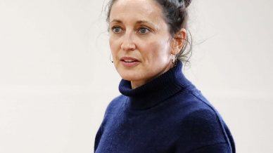 A woman with fair skin and brown hair in a messy bun is wearing a navy blue polo-neck jumper, standing against a plain light background, looking slightly to the side.