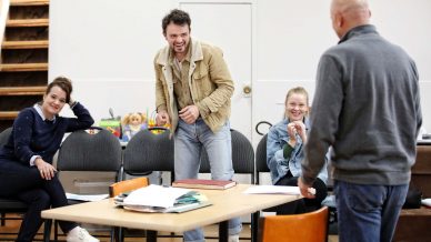 Four people are in a casual, cheerful meeting room. Three are seated smiling or laughing, whilst one man stands, also laughing. Papers and folders are on a table in the foreground, with shelves and toys in the background.