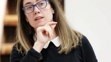 A woman with long brown hair and glasses, wearing a black top with a white collar, gestures with her hand near her chin whilst speaking. She stands indoors in front of a blurred background.