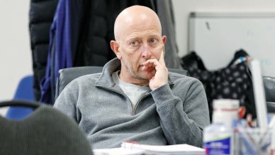A bald man in a grey jumper sits at a cluttered desk, resting his chin on his hand and looking thoughtful. Coats hang in the background and office supplies are scattered on the desk.