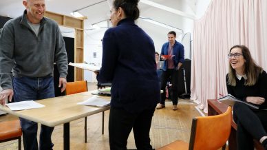 Four adults rehearse in a bright room. One man stands by a table, smiling. A woman stands nearby with her back to the camera. Two others sit and laugh, holding scripts. The mood is relaxed and cheerful.