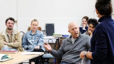 A man sits in a classroom, gesturing expressively towards a woman standing in front of him. Several people sit and watch the interaction, appearing attentive and engaged. Books and papers are on the tables around them.
