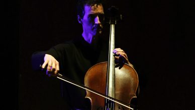A musician in dark clothing plays a cello onstage under dramatic lighting, highlighting the instrument and the musician’s face against a dark background.