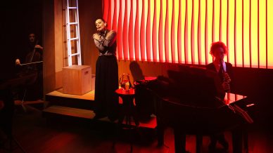 A woman stands on stage with hands clasped near her face, a cellist sits to the left, and a pianist sits at a grand piano. The background features a bright, glowing orange and red panel.