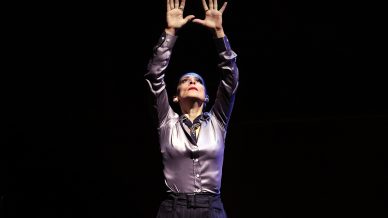 A person wearing a shiny silver shirt and dark trousers stands on stage, raising both hands above their head with fingers spread wide, looking upward under dramatic lighting.