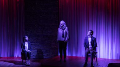 Three people under dramatic purple and red stage lighting; one stands on a platform in the centre, while the other two sit on chairs on either side, against a backdrop of curtains and a stone wall.
