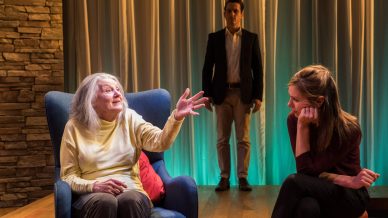 An elderly woman gestures while sitting in a blue armchair, speaking to a young woman seated nearby. A man in a suit stands in the background. The scene is warmly lit with stone and curtain backdrops.