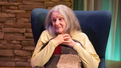 An elderly woman with long grey hair sits in a blue armchair, hands clasped to her chest, wrapped in a patchwork blanket. She wears a yellow jumper and has a warm, gentle expression. A stone wall is in the background.