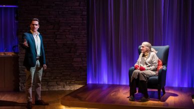 A man in a blue blazer stands gesturing with his hand, while an elderly woman with grey hair sits in an armchair on a lit wooden platform, with dramatic purple lighting and a stone wall in the background.