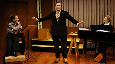 A man stands centre stage singing with his arms outstretched, while a woman on the left sits with a score and another woman on the right plays the piano. All are dressed in formal attire in a wood-panelled music room.