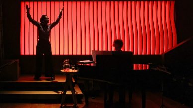 A person sings with raised arms beside a pianist on stage, both silhouetted against a dramatic, wavy red-lit backdrop. A table with a glass jug and glass sits next to the piano.