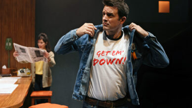 A man stands adjusting his denim jacket to reveal a T-shirt with the words “GET ‘EM DOWN!” printed in red. In the background, a woman reads a newspaper at a kitchen worktop. The setting appears to be a staged scene.