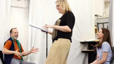Three adults talk and laugh together indoors. One man sits gesturing, while a standing woman holds papers and smiles. Another woman, seated, listens and smiles. The background has white curtains and technical equipment.