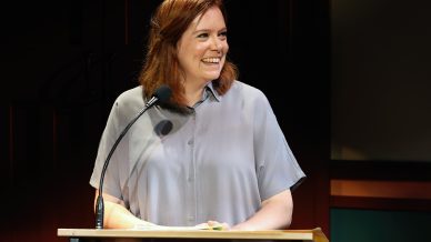 A woman with red hair and a light purple blouse smiles whilst standing at a lectern with a microphone, speaking in a warmly lit indoor setting.