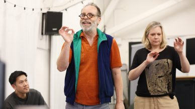 Three adults are indoors, engaged in discussion. A man in an orange shirt and blue waistcoat gestures while smiling; a woman in a grey shirt gestures beside him, and another man sits nearby, listening. The setting appears casual and collaborative.