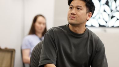 A man in a dark T-shirt sits on a chair, looking thoughtfully to the side. In the background, a blurred person is seated and a decorative pattern is visible on the wall.