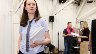 A woman in a light blue shirt and loose necktie holds scripts, appearing thoughtful in a rehearsal space. In the background, a man and woman talk, holding papers. The room has white walls and exposed beams.