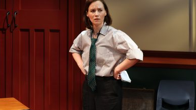 A woman wearing a striped shirt, loose tie, and dark trousers stands with hands on her hips, looking concerned. She holds papers in one hand and stands beside a desk and an empty chair in a room with red panelling.