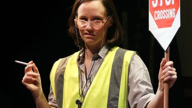 A woman in a yellow safety vest holds a Children Stop Crossing sign in one hand and a cigarette in the other, standing against a dark background.