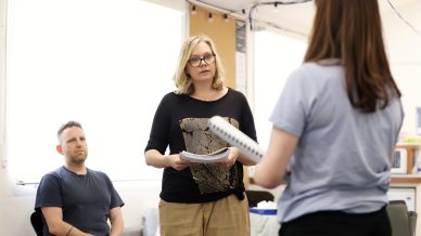 A woman holding papers stands and speaks to another woman with her back to the camera, whilst a seated man listens in a brightly lit room.