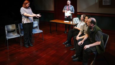 A woman stands and gestures whilst speaking to three seated people in casual clothing. Another person stands behind them holding papers. The setting is a room with wooden floors and plastic chairs.