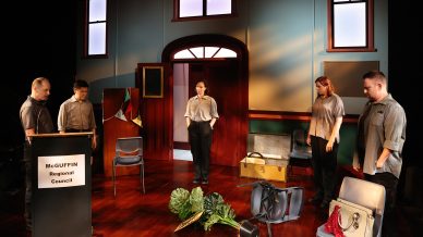 Five people stand in a council room set with scattered chairs and luggage, a tipped-over plant, and a lectern labelled McGUFFIN Regional Council. The mood appears tense or sombre.