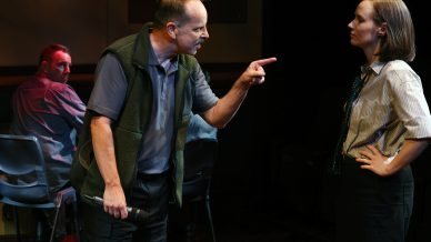 A man stands pointing and speaking angrily to a woman with her hands on her hips, whilst another man in the background looks over his shoulder at them. The scene appears tense and dramatic, set indoors with subdued lighting.