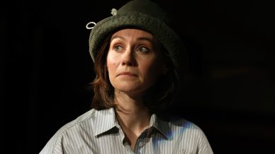 A woman wearing a striped shirt and a green bucket hat looks slightly upwards with a thoughtful expression, illuminated against a dark background.