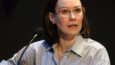 A woman with shoulder-length brown hair and glasses speaks at a lectern with a microphone, wearing a striped button-up shirt. She appears engaged and expressive, with one arm resting on the lectern.