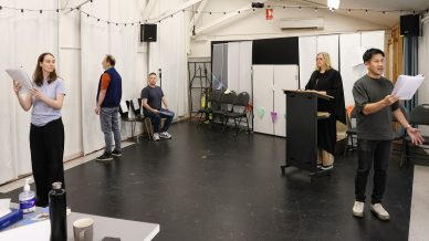 Five people rehearse a script in a spacious room with white walls. Two hold scripts and stand apart, one sits on a chair, one faces the wall, and another stands at a lectern. Empty chairs and fairy lights decorate the background.