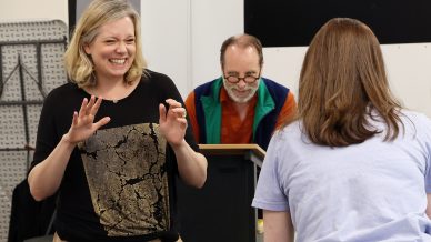 A woman smiles and gestures animatedly towards another person with their back to the camera, whilst an older man reads from a lectern in the background. The scene appears to be a lively rehearsal or group activity.