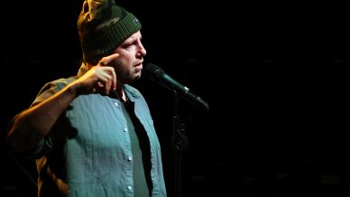 A man wearing a camouflage beanie and blue shirt speaks passionately into a microphone on a dark stage, illuminated by a spotlight.