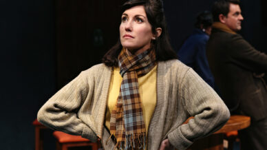 A woman in a beige cardigan and checked scarf stands with hands on hips, looking upward thoughtfully. A man in a brown suit sits in the background, facing away. The setting appears to be a stage play.