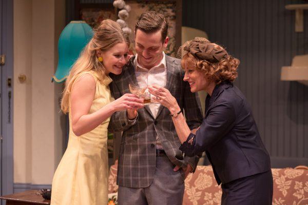 Three people dressed in vintage clothing stand closely together in a living room, smiling and holding glasses in a toast. The room features retro decor, including a blue lamp and patterned sofa.