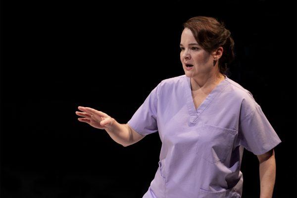 A woman wearing light purple medical scrubs gestures with her right hand, extending her fingers as she stands against a dark background. She appears to be speaking or explaining something.