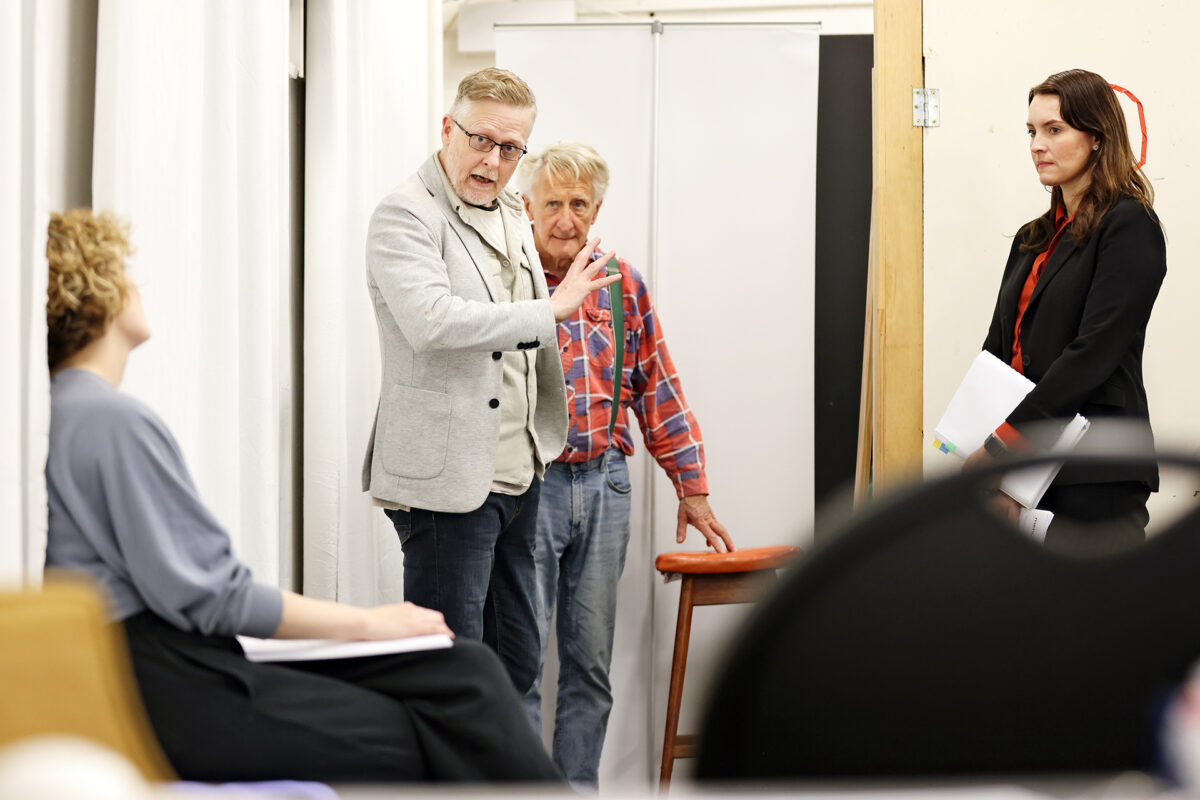 A man gestures while speaking to a seated woman, as another older man and a woman holding papers watch attentively in a rehearsal or meeting room.