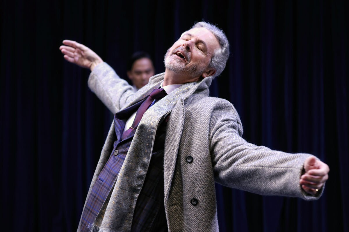 A man with grey hair and a beard, wearing a coat and suit, stands with arms outstretched and eyes closed on stage, conveying emotion. Another person is blurred in the background against a dark curtain.