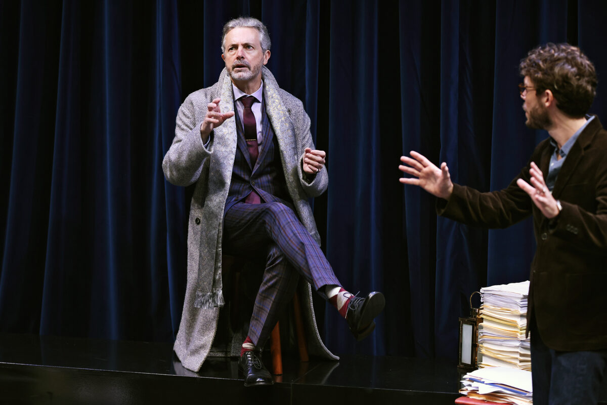A man in a grey coat sits on a stool, gesturing with one hand, in front of dark blue curtains. Another man stands nearby with arms raised, next to a stack of papers and a lantern. Both appear to be in conversation on stage.