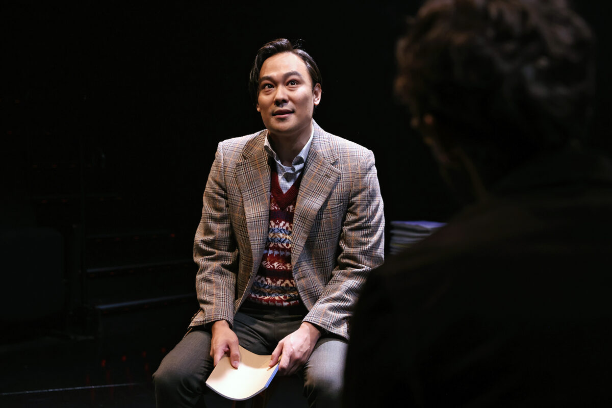 A man in a checked blazer and patterned jumper vest sits onstage holding a sheet of paper, looking up attentively at a person with their back to the camera in a dimly lit theatre setting.