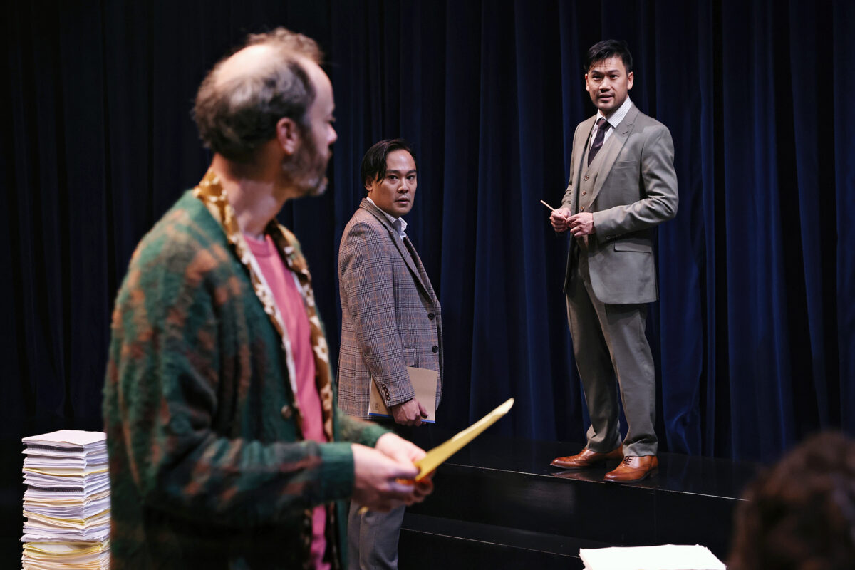 Three men in formal and semi-formal attire are on a stage with dark curtains; one stands elevated holding papers, while two others, one with a beard, look towards him. A stack of papers sits in the foreground.