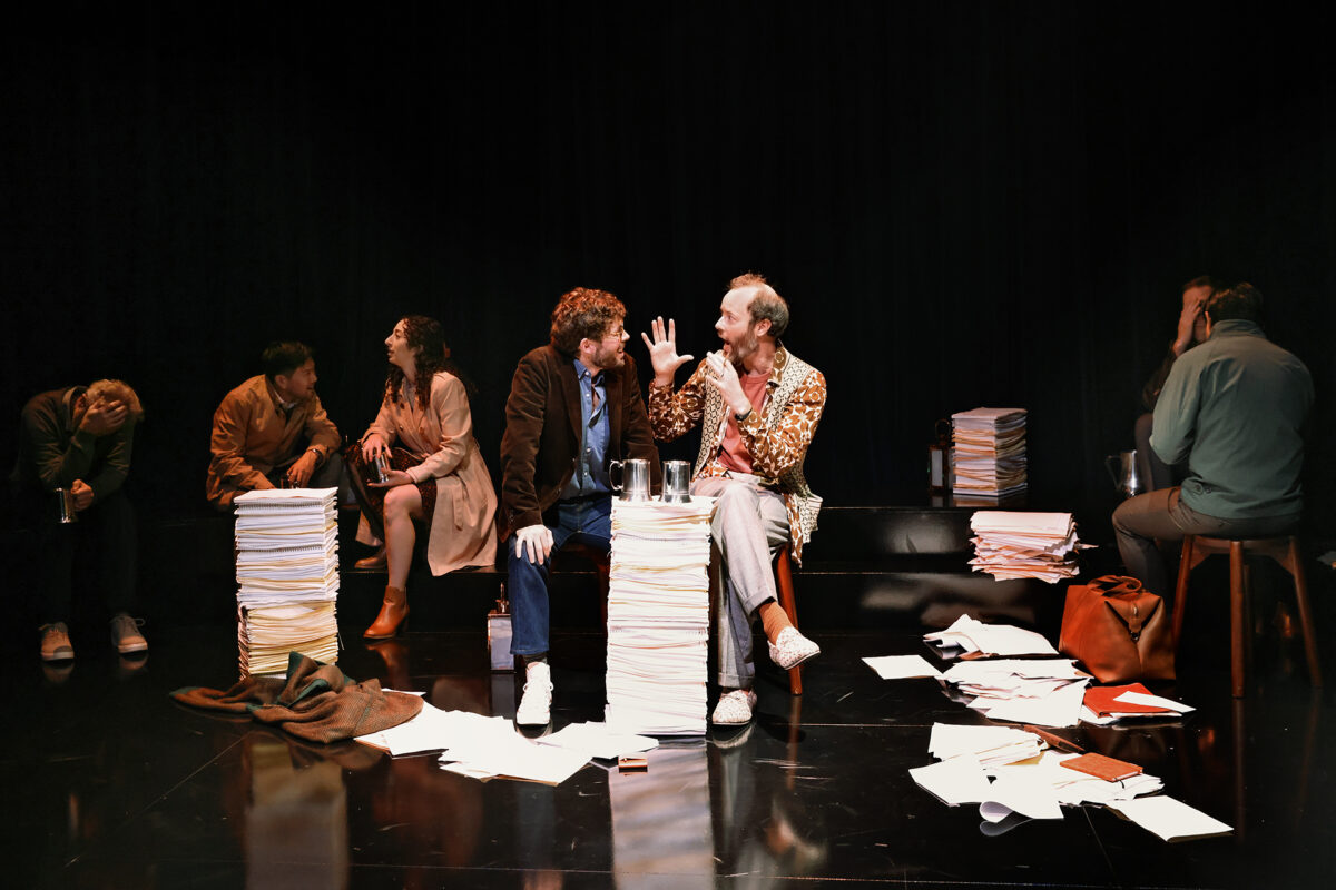 Seven people sit on a stage among stacks of papers, some talking animatedly and others looking distressed. Loose papers are scattered on the floor, and the scene appears tense and dramatic.