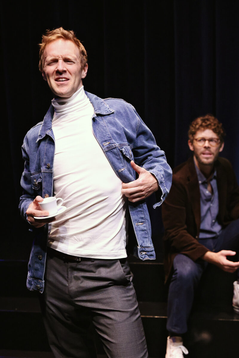 A man in a white polo neck and denim jacket holds a small cup and saucer, posing confidently. Another man in glasses and a brown jacket sits in the background against a dark curtain.