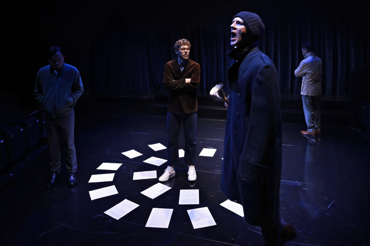 Four actors on a dimly lit stage; one stands at the centre among scattered papers, another shouts in the foreground, and two others stand apart, facing away, creating a tense atmosphere.