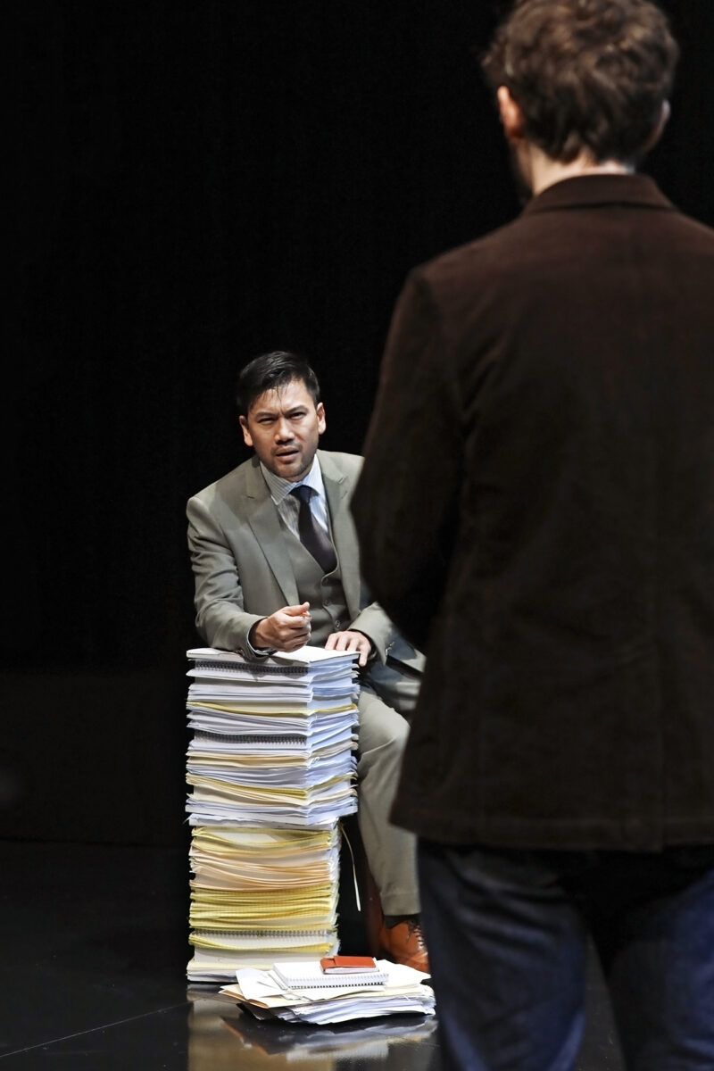 A man in a suit sits on a tall stack of papers, speaking with a serious expression to another person standing with their back to the camera, against a dark background.