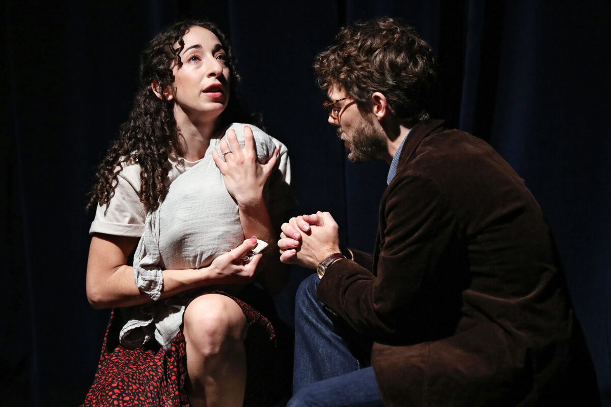 A woman clutching a cloth looks up emotionally while seated next to a bearded man in glasses and a brown jacket; they appear to be having an intense conversation on stage with a dark curtain backdrop.