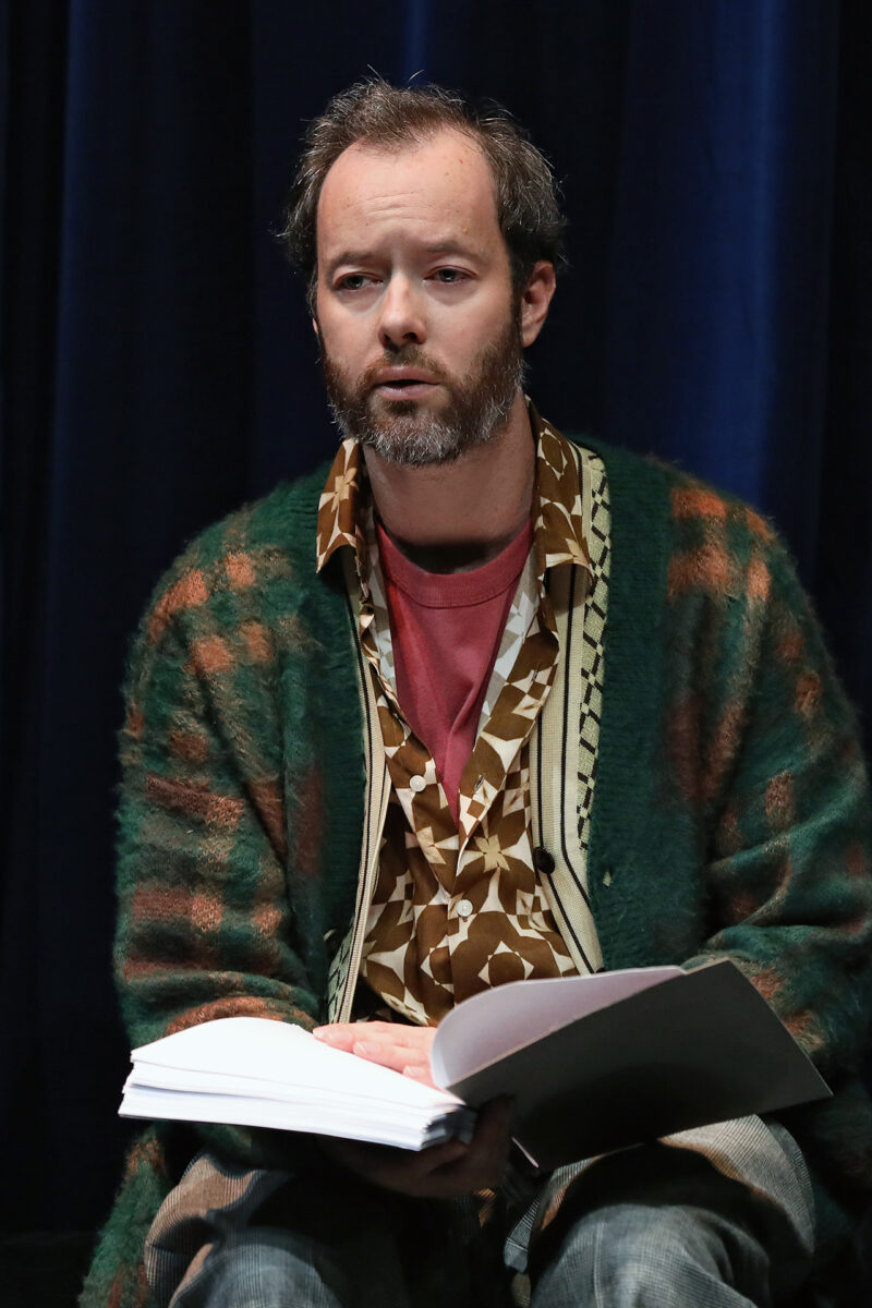 A man with a beard sits in front of dark blue curtains, wearing a patterned shirt and a green cardigan, holding an open script or book in his lap and looking slightly to the side.